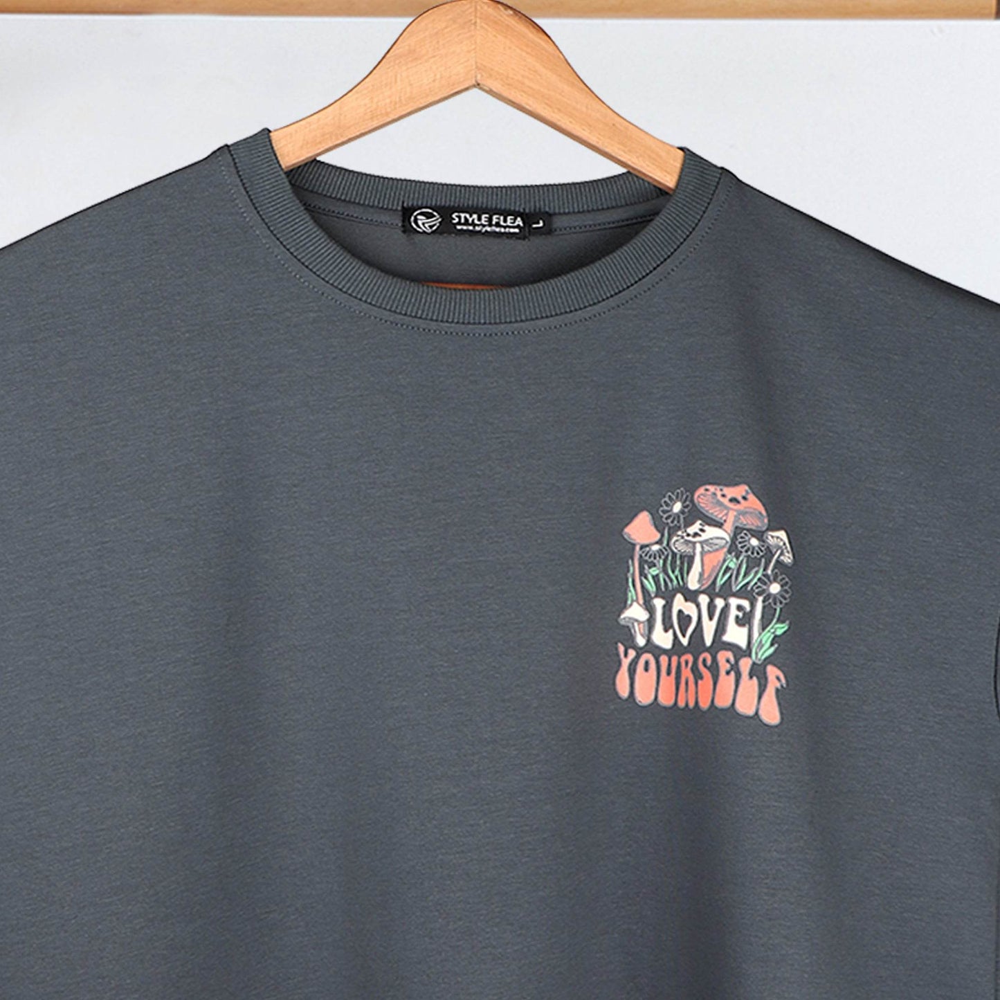 Close-up of a charcoal blue drop shoulder T-shirt on a hanger, featuring a small "Love Yourself" mushroom and floral graphic on the chest.