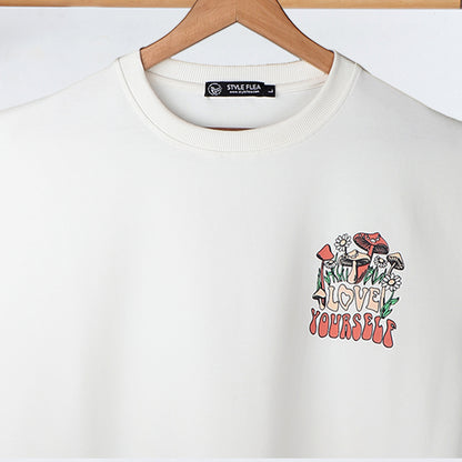 Close-up of a white drop shoulder T-shirt on a hanger, featuring a small "Love Yourself" mushroom and floral graphic on the chest.